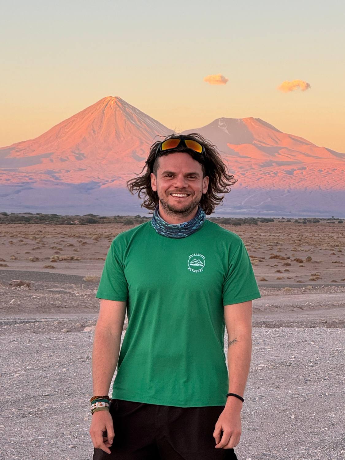 Retrato de Erick sonriendo en el desierto, con camiseta verde y volcanes al atardecer detrás